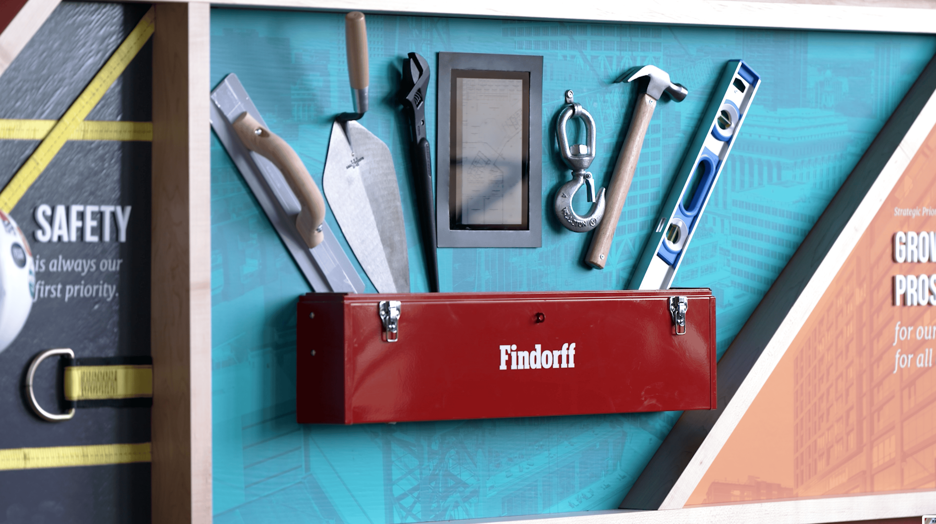 Display of construction tools and equipment in a branded Findorff toolbox setup by Thysse. The installation features a trowel, wrench, hammer, and level, mounted against a blueprint-themed backdrop as part of the facility branding project.